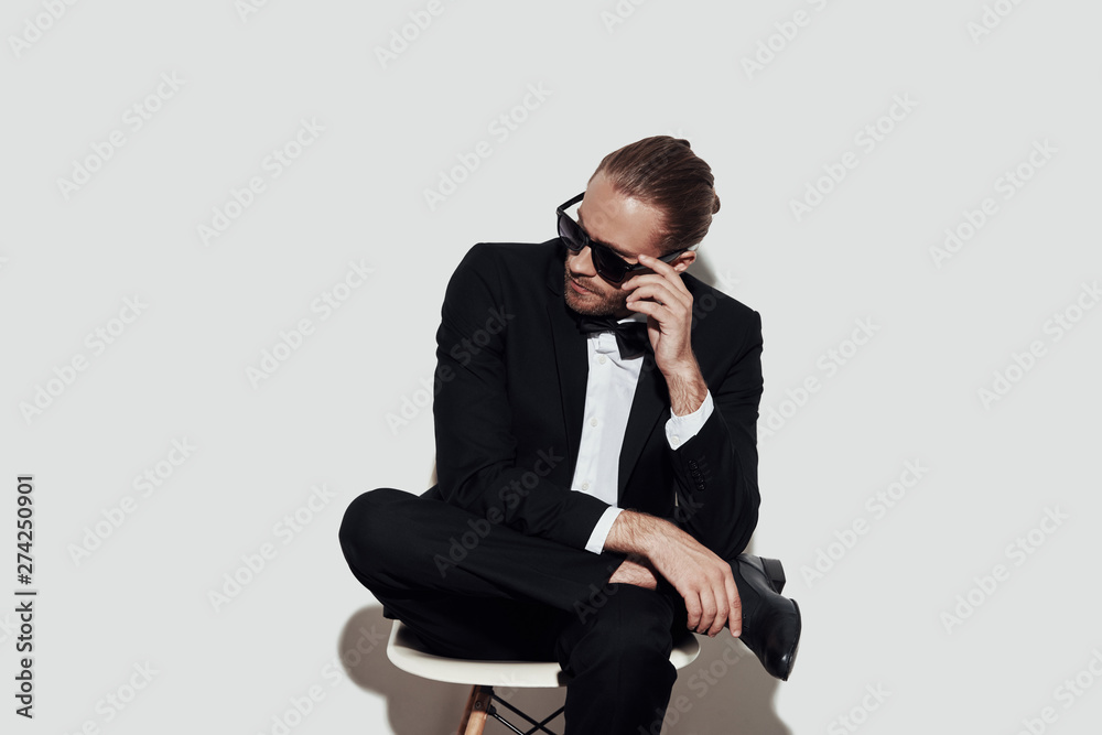He got the look. Charming young man in full suit looking away and adjusting sunglasses while sitting against grey background He got the look. Charming young man in full suit looking away and adjusting sunglasses while sitting against grey background