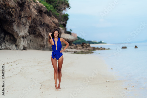 Beautiful sexy tanned woman in blue swimsuit on white sand beach 