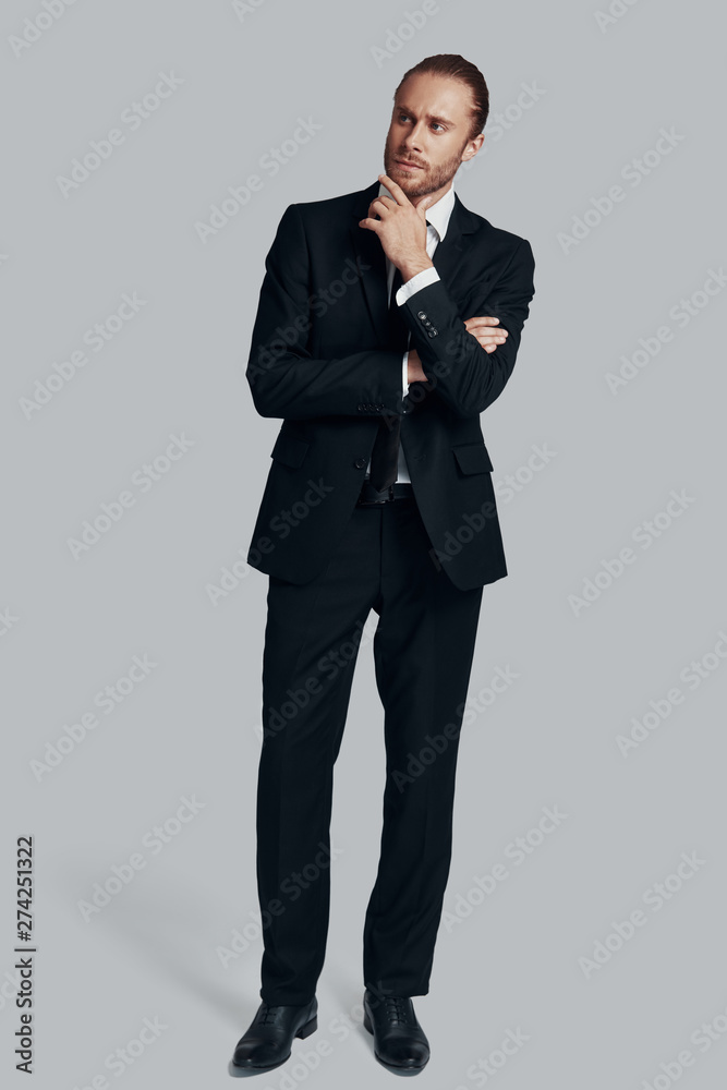 Contemplation. Full length of young man in full suit looking away while standing against grey background Contemplation. Full length of young man in full suit looking away while standing against grey background