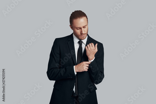 Portrait of confidence. Handsome young man in full suit adjusting his sleeve while standing against grey background