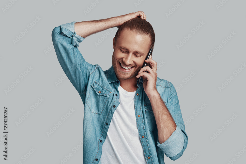 Pleasant talk. Handsome young man talking on smart phone and smiling while standing against grey background Pleasant talk. Handsome young man talking on smart phone and smiling while standing against grey background