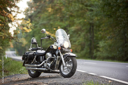 Schilderij op canvas Black shiny powerful cruiser motorcycle parked on roadside on background of green trees on sunny evening