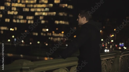 Depressed handsome man with beard and brown hair leaning on a railing of the bridge, thinking sadly to his ex girlfriend on blurred city colorful lights background. Medium Shot. photo