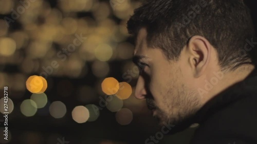 Sad handsome man with beard and brown hair leaning on a railing of the bridge, thinking sadly to his ex girlfriend on blurred city colorful lights background.