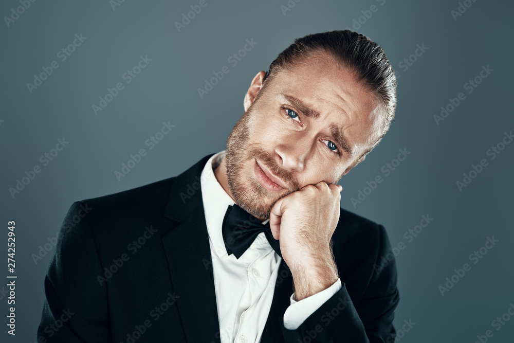 Bored. Charming young man in full suit leaning on his hand and making a face while standing against grey background Bored. Charming young man in full suit leaning on his hand and making a face while standing against grey background