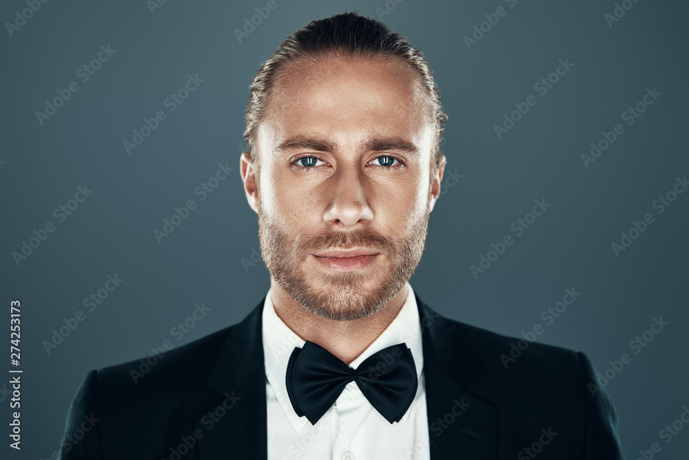 Confident look. Charming young man in bow tie looking at camera and smiling while standing against grey background Confident look. Charming young man in bow tie looking at camera and smiling while standing against grey background