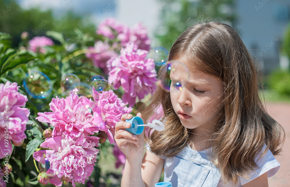 Happy little child blowing soap bubbles outdoors in summer park. Blooming pink peony (Paeonia, peony) flower. Enjoy nature. Healthy girl on spring lawn. Allergy free concept. Freedom