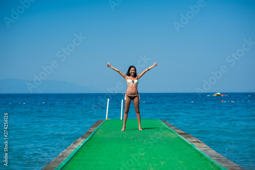 slender young girl stands on the pier, Mediterranean Sea, azure water, sunny, tanned skin, brunette with long hair, green pier, sunbathes, tropical vacation, relax, have fun under the rays © SYARGEENKA
