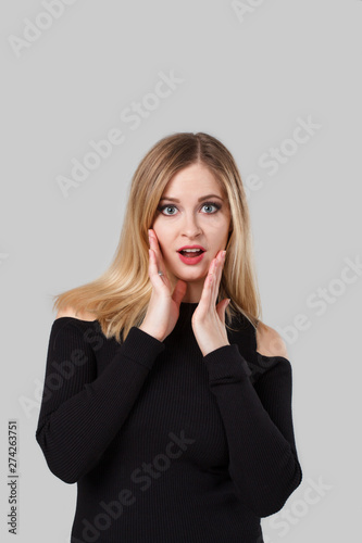 Blond girl is surprised and is standing in front of a gray background. She is wearing a black sweater.