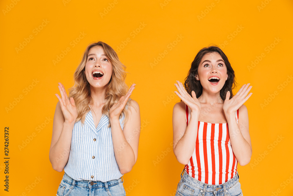 Portrait of two beautiful blonde and brunette women 20s in summer wear expressing delight while raising arms Portrait of two beautiful blonde and brunette women 20s in summer wear expressing delight while raising arms