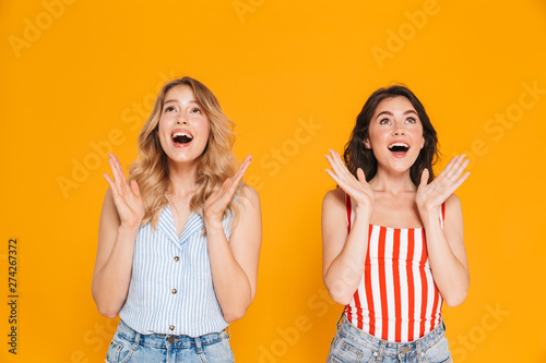 Portrait of two beautiful blonde and brunette women 20s in summer wear expressing delight while raising arms