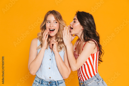 Portrait of two pretty blonde and brunette women 20s in summer wear expressing surprise while whispering gossips photo