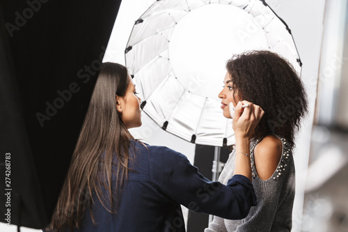 Portrait of female makeup artist applying professional cosmetics to gorgeous woman during photo shooting in studio