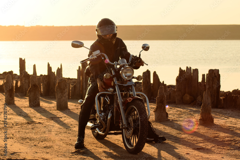 Obraz premium biker in the leather and helm on the red motorcycle against field and seashore. Brutal man in leather at sunset