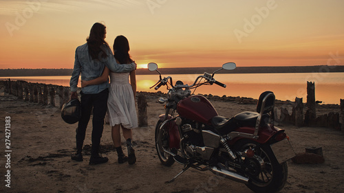 Romantic biker couple with red motorcycle. Handsome bearded long-haired man and attractive woman outdoors against motorcycle and sunset, sunrise