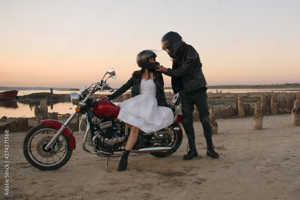 Obraz premium Romantic biker couple in leather and helms with red motorcycle. Woman and man against motorcycle and sunset, sunrise