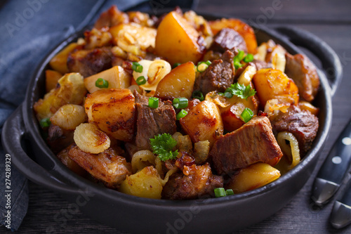 Fried beef and  potatoes with onions and garlic served in black dish on wooden background