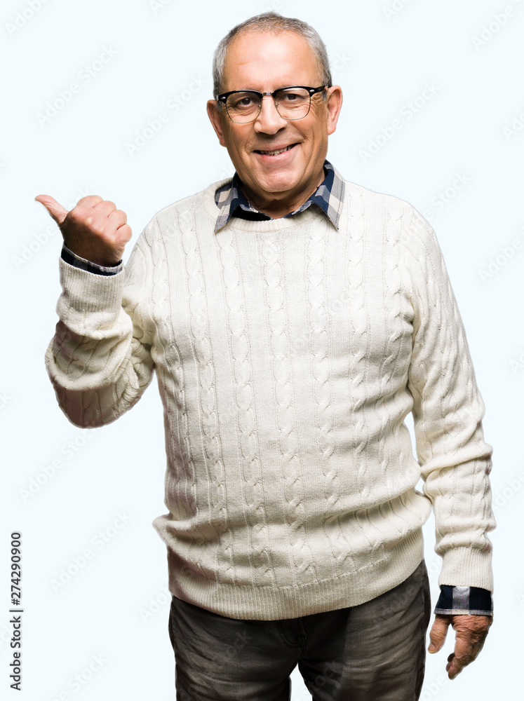 Handsome senior man wearing glasses and winter sweater smiling with happy face looking and pointing to the side with thumb up. Handsome senior man wearing glasses and winter sweater smiling with happy face looking and pointing to the side with thumb up.