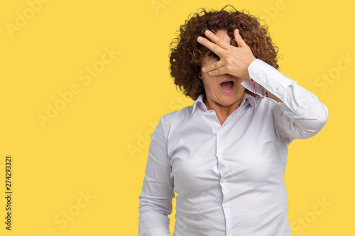 Beautiful middle ager senior businees woman over isolated background peeking in shock covering face and eyes with hand, looking through fingers with embarrassed expression. © Krakenimages.com