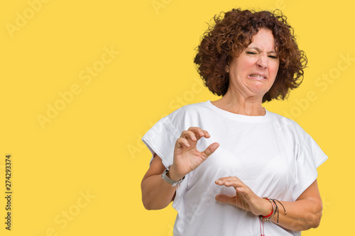 Beautiful middle ager senior woman wearing white t-shirt over isolated background disgusted expression, displeased and fearful doing disgust face because aversion reaction. With hands raised