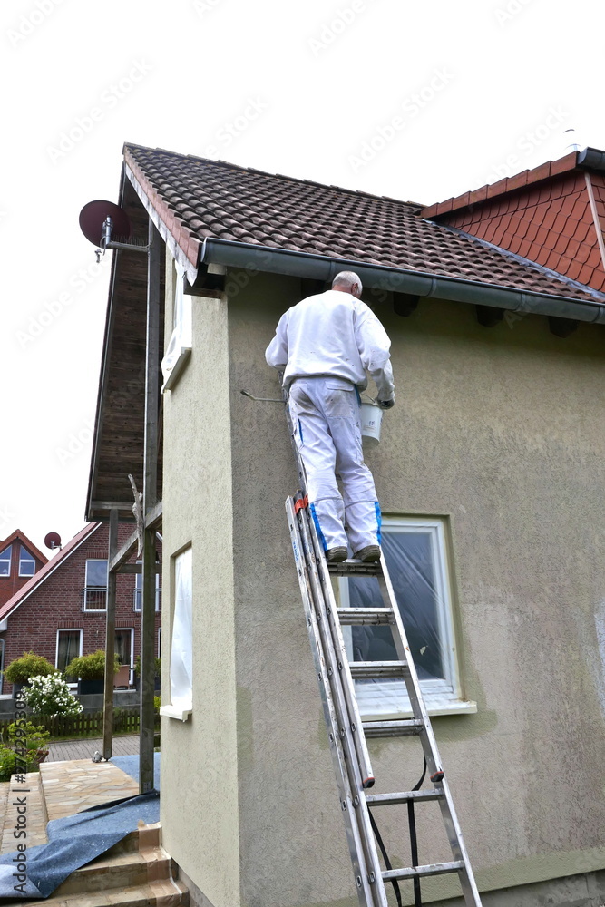 Rear view of a painter standing on a ladder and stroking the outside wall of a house with a paint roller. Rear view of a painter standing on a ladder and stroking the outside wall of a house with a paint roller.