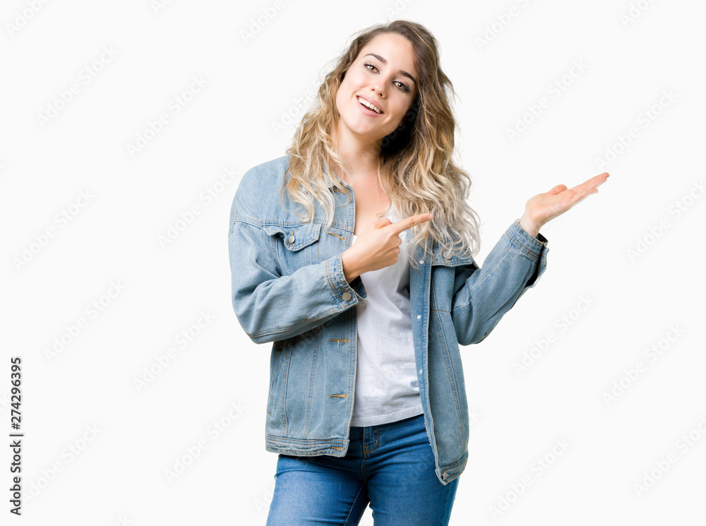Beautiful young blonde woman wearing denim jacket over isolated background amazed and smiling to the camera while presenting with hand and pointing with finger. Beautiful young blonde woman wearing denim jacket over isolated background amazed and smiling to the camera while presenting with hand and pointing with finger.