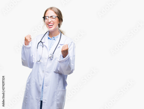 Beautiful young blonde doctor woman wearing medical uniform over isolated background very happy and excited doing winner gesture with arms raised, smiling and screaming for success