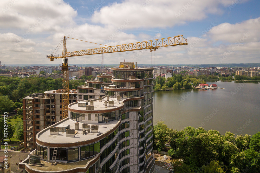Obraz premium Apartment or office tall building unfinished under construction among green tree tops. Tower cranes on bright blue sky copy space background.