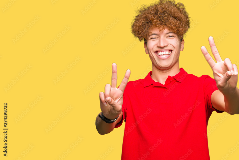 Young handsome man with afro hair wearing red t-shirt smiling looking to the camera showing fingers doing victory sign. Number two.