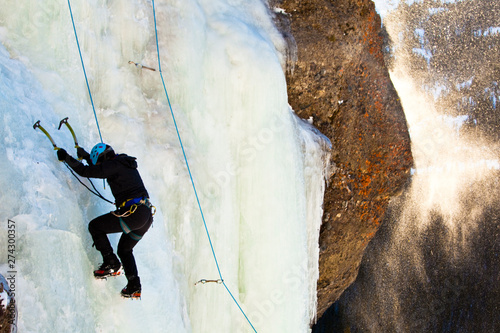 A woman climbs Over Easy (WI3) at Hyalite Canyon in Montana as spindrift swirls by in the afternoon sunlight.