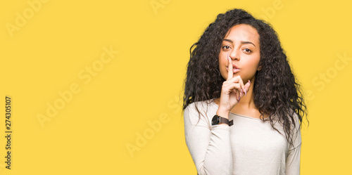Young beautiful girl with curly hair wearing casual sweater asking to be quiet with finger on lips. Silence and secret concept.