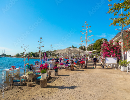 Fototapeta Naklejka Na Ścianę i Meble -  View of restaurant or cafe and bougainvillea flowers on beach in Gumusluk, Bodrum city of Turkey. Aegean seaside style colorful chairs, tables and flowers in Bodrum town near beautiful Aegean Sea.