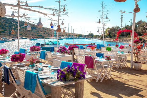 Fototapeta Naklejka Na Ścianę i Meble -  View of restaurant or cafe and bougainvillea flowers on beach in Gumusluk, Bodrum city of Turkey. Aegean seaside style colorful chairs, tables and flowers in Bodrum town near beautiful Aegean Sea.