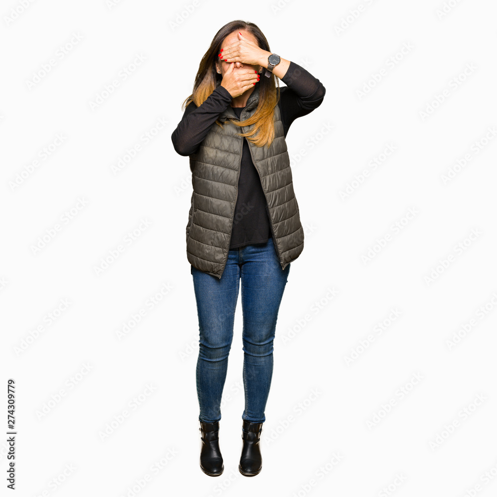 Beautiful middle age woman wearing winter vest Covering eyes and mouth with hands, surprised and shocked. Hiding emotion Beautiful middle age woman wearing winter vest Covering eyes and mouth with hands, surprised and shocked. Hiding emotion