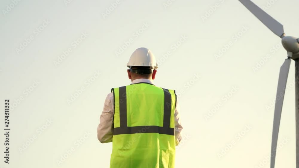 Engineer in hard hat and bright vest is near the windmill and watching to see how it works