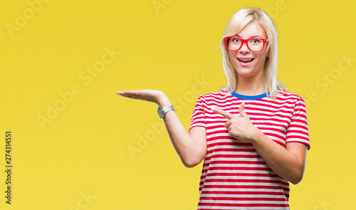Young beautiful blonde woman wearing glasses over isolated background amazed and smiling to the camera while presenting with hand and pointing with finger.