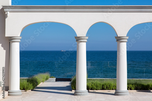 an arch with columns of white stone and a marble floor with a lawn and a view of the sea on the coast of a luxurious mansion.