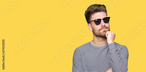 Young handsome man wearing sunglasses with hand on chin thinking about question, pensive expression. Smiling with thoughtful face. Doubt concept.