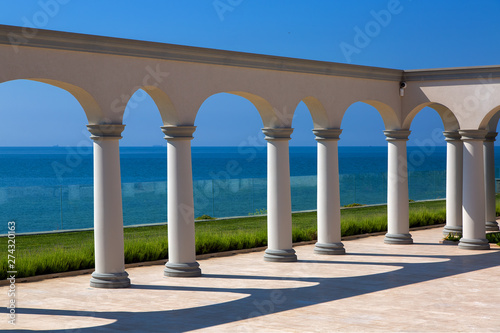 an arch with columns of white stone on sunny day and a marble floor with a lawn and a view of the sea on the coast of a luxurious mansion.