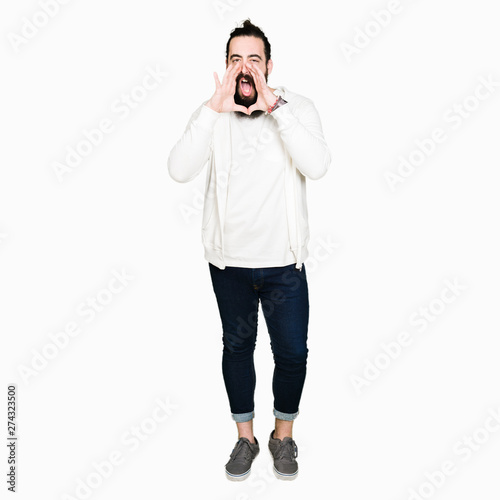 Young hipster man with long hair and beard wearing sporty sweatshirt Shouting angry out loud with hands over mouth