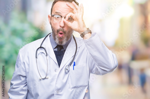 Middle age senior hoary doctor man wearing medical uniform isolated background doing ok gesture shocked with surprised face, eye looking through fingers. Unbelieving expression.