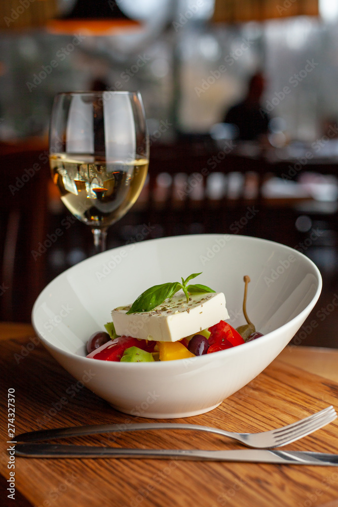Traditional fresh greek salad with cherry tomatoes, bell pepper, cucumber and feta cheese dressed with olive oil served with white wine on wooden table with blurred background