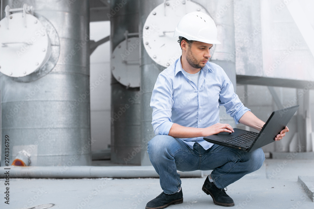 Young engineer in hardhat sitting on the background of water or gas pipes station and using laptop. Heating station manager doing his job outdoor. Young engineer in hardhat sitting on the background of water or gas pipes station and using laptop. Heating station manager doing his job outdoor.