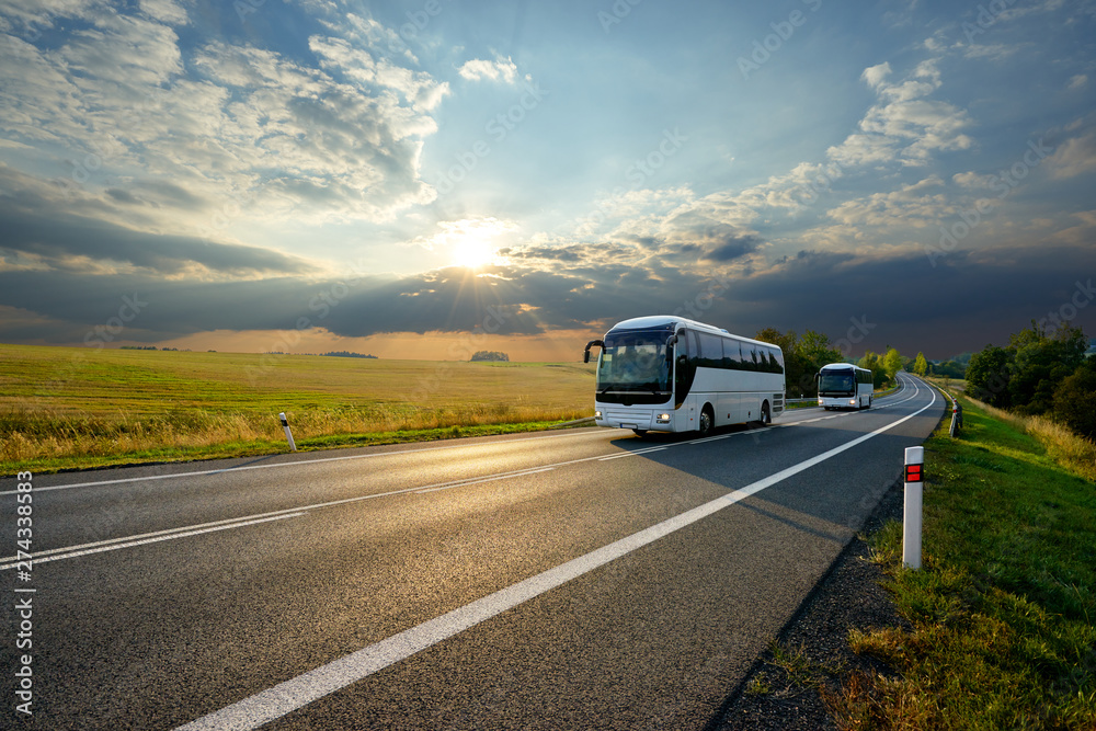 Two white buses traveling on the asphalt road in rural landscape at sunset with dramatic clouds