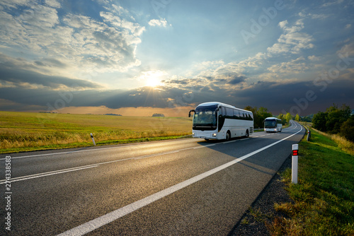 Two white buses traveling on the asphalt road in rural landscape at sunset with dramatic clouds photo