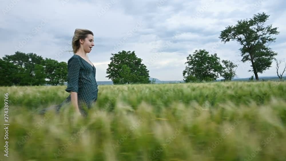 Young happy blonde girl runs on a green wheat field in the evening against the background of the rain sky. Side view Young happy blonde girl runs on a green wheat field in the evening against the background of the rain sky. Side view