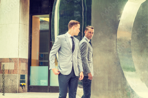 Young Businessman Fashion in New York City. Young Man wearing gray blazer, white shirt, black tie, black pants, standing on street in front of metal mirror, looking at reflections, thinking..