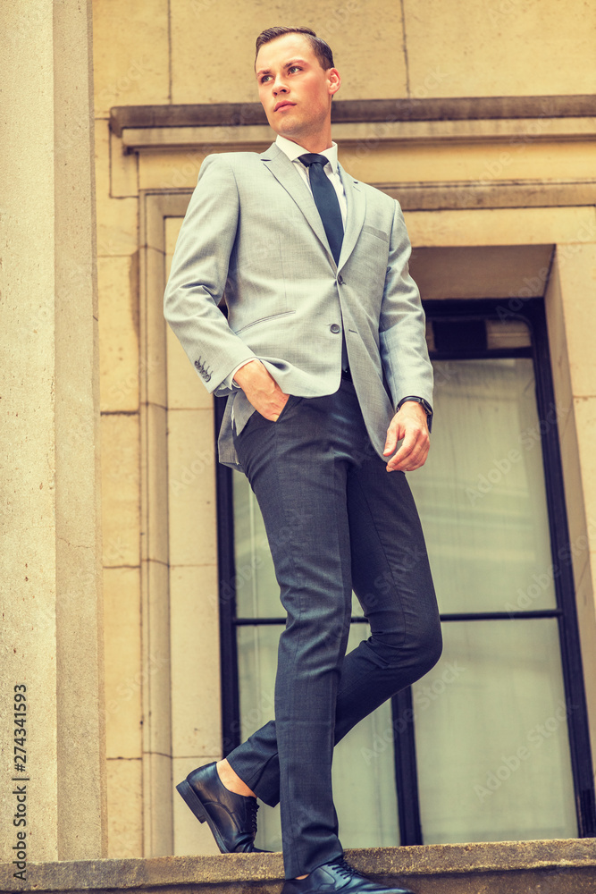 Young Businessman Street Fashion in New York City. Young Man wearing gray blazer, white shirt, black tie, black pants, leather shoes, walking down stairs by window outside office building, looking. Young Businessman Street Fashion in New York City. Young Man wearing gray blazer, white shirt, black tie, black pants, leather shoes, walking down stairs by window outside office building, looking.