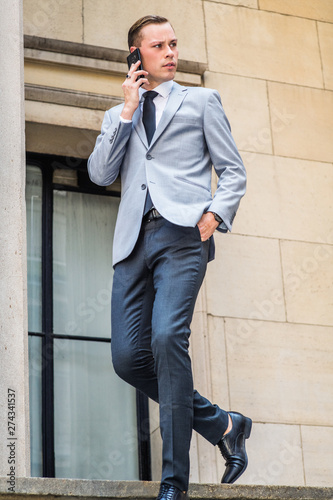Young Businessman Street Fashion in New York City. Man wearing gray blazer, white shirt, black tie, pants, leather shoes, walking down stairs by window outside office building, talking on cell phone..