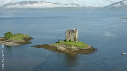 An aerial view of Castle Stalker on Loch Laich on a sunny morning. Flying right to left whilst panning left to right and zooming in, with mountains and low clouds behind the castle. photo